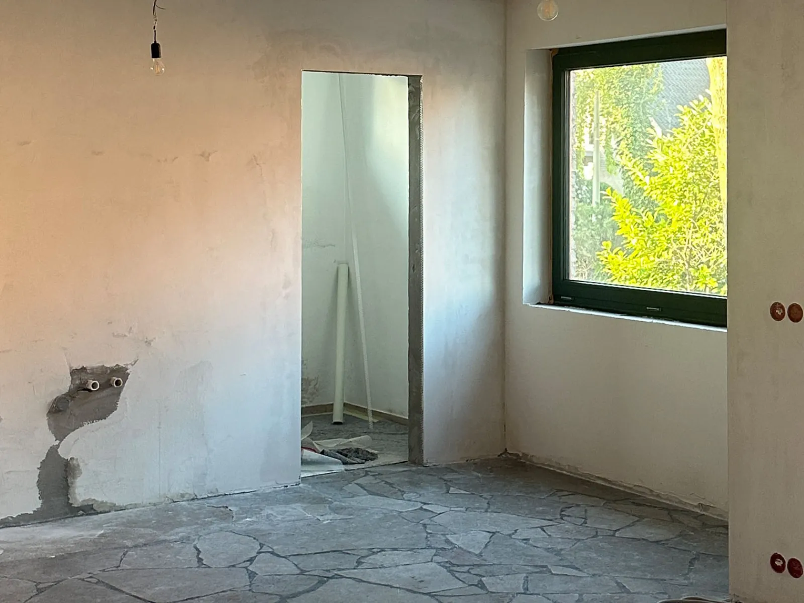 Empty room under construction with stone tile floor, unfinished walls, exposed wiring, and a large window showing green foliage outside.