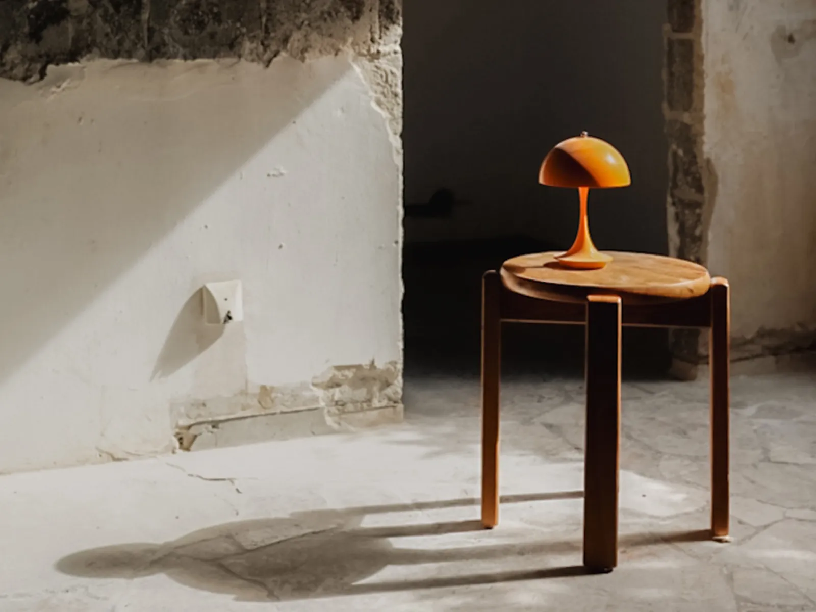 Wooden stool with a small orange lamp on top, casting shadows on a cracked concrete floor and worn white wall.