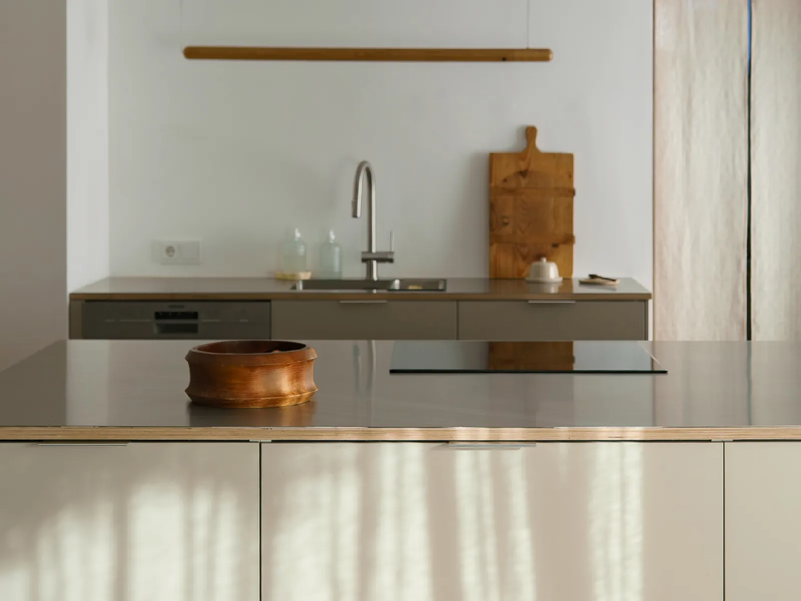Minimalist kitchen with a wooden bowl on a gray island, modern faucet, wooden cutting board, and soap dispensers against a white wall.