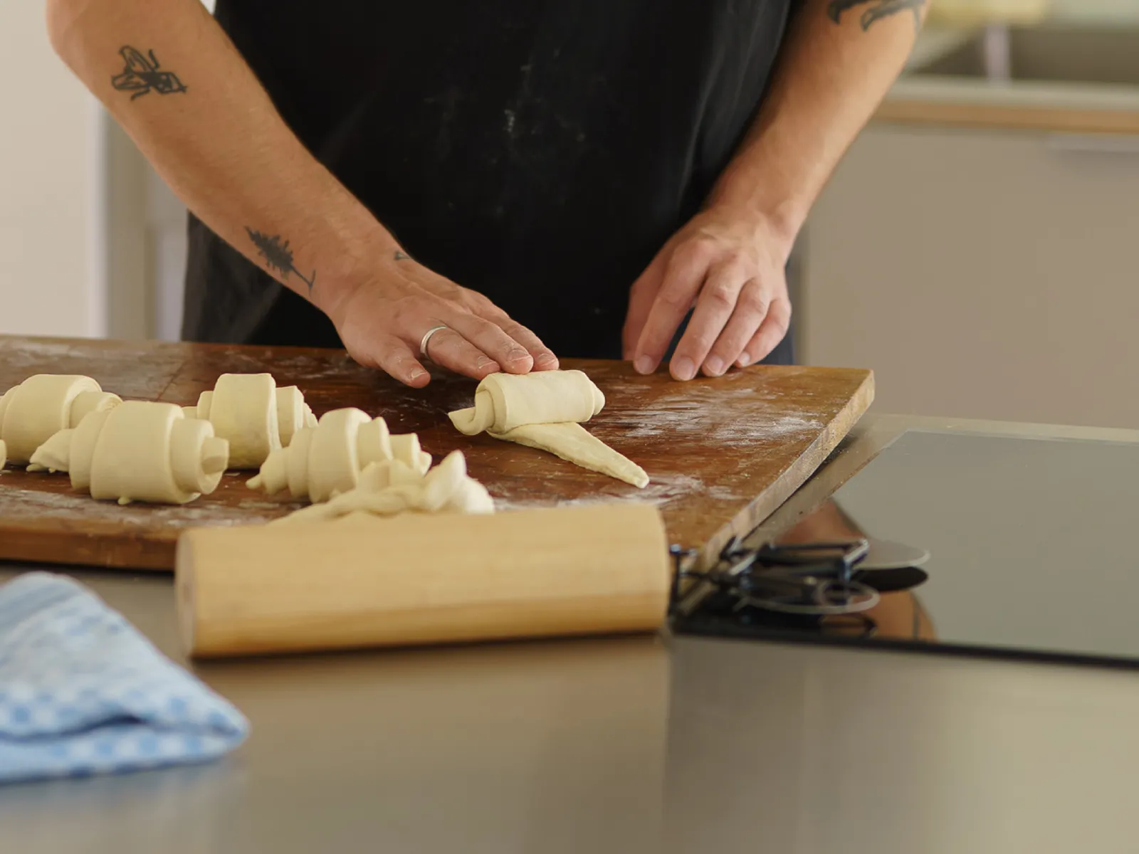 Hands with tattoos are rolling croissant dough on a wooden board next to a rolling pin and an induction hob.
