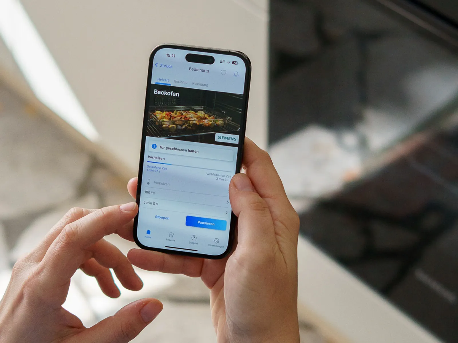 Person using a smartphone app to remotely control a Siemens oven baking food at 180°C, with options to pause or stop.