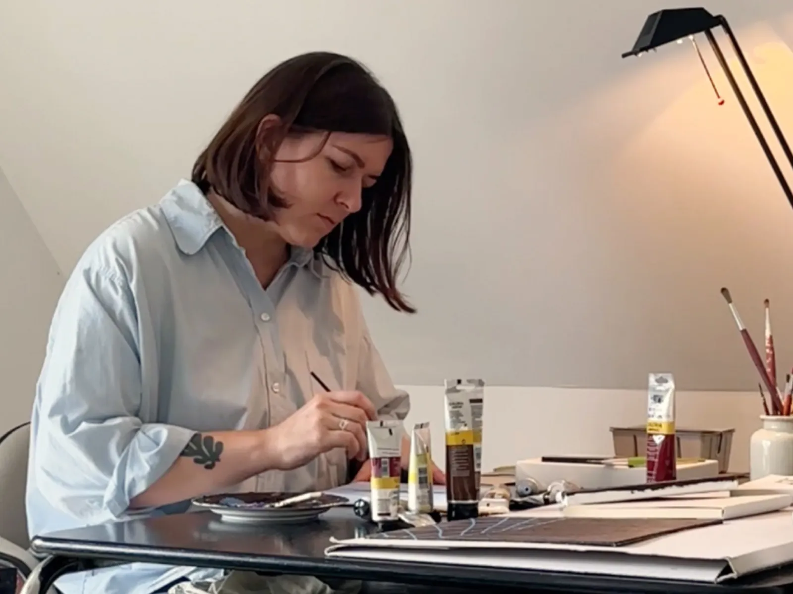 Person in a light blue shirt painting with brushes and tubes of paint at a table in a well-lit room.