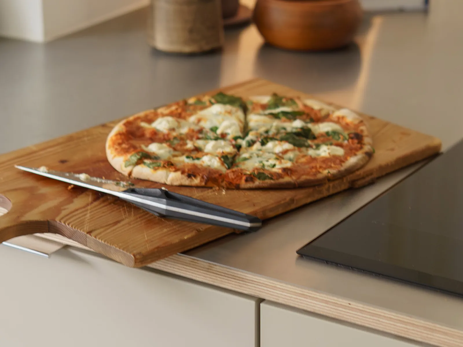 Wooden cutting board with sliced pizza and a knife on a kitchen countertop next to a stovetop.