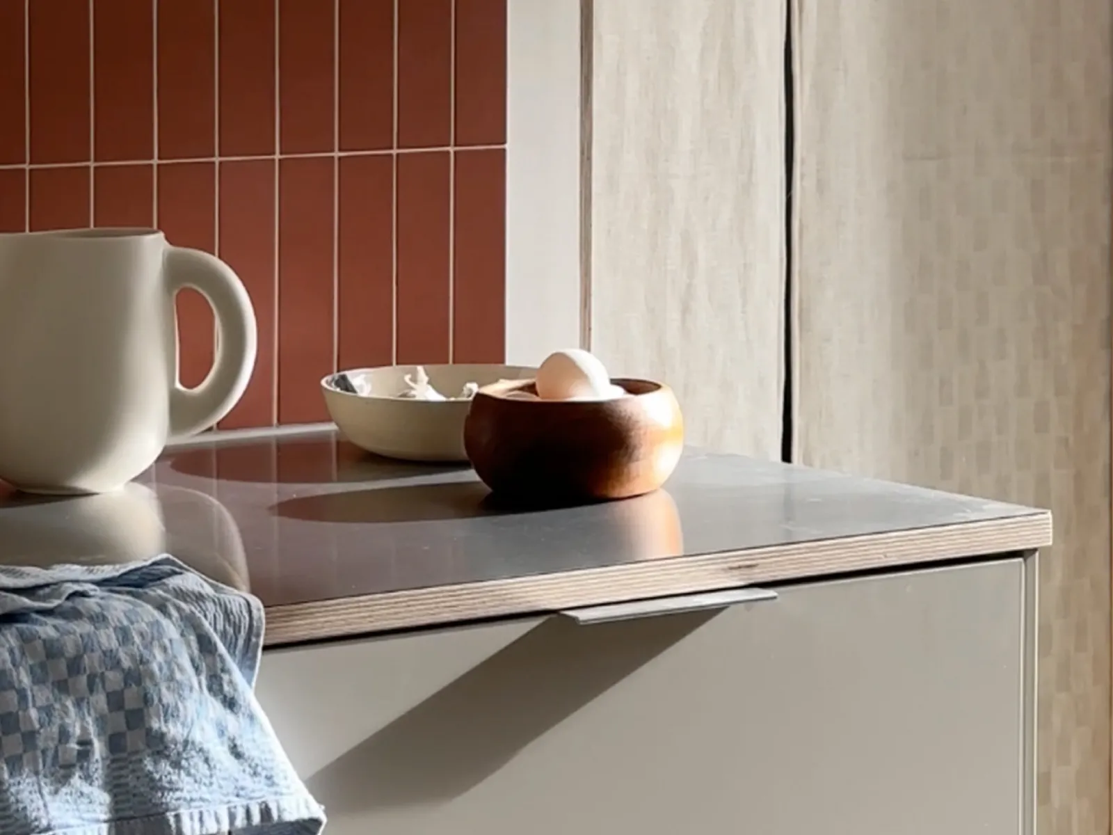 Minimalist kitchen counter with a ceramic pitcher, wooden and ceramic bowls, and a blue checkered cloth draped over the edge.