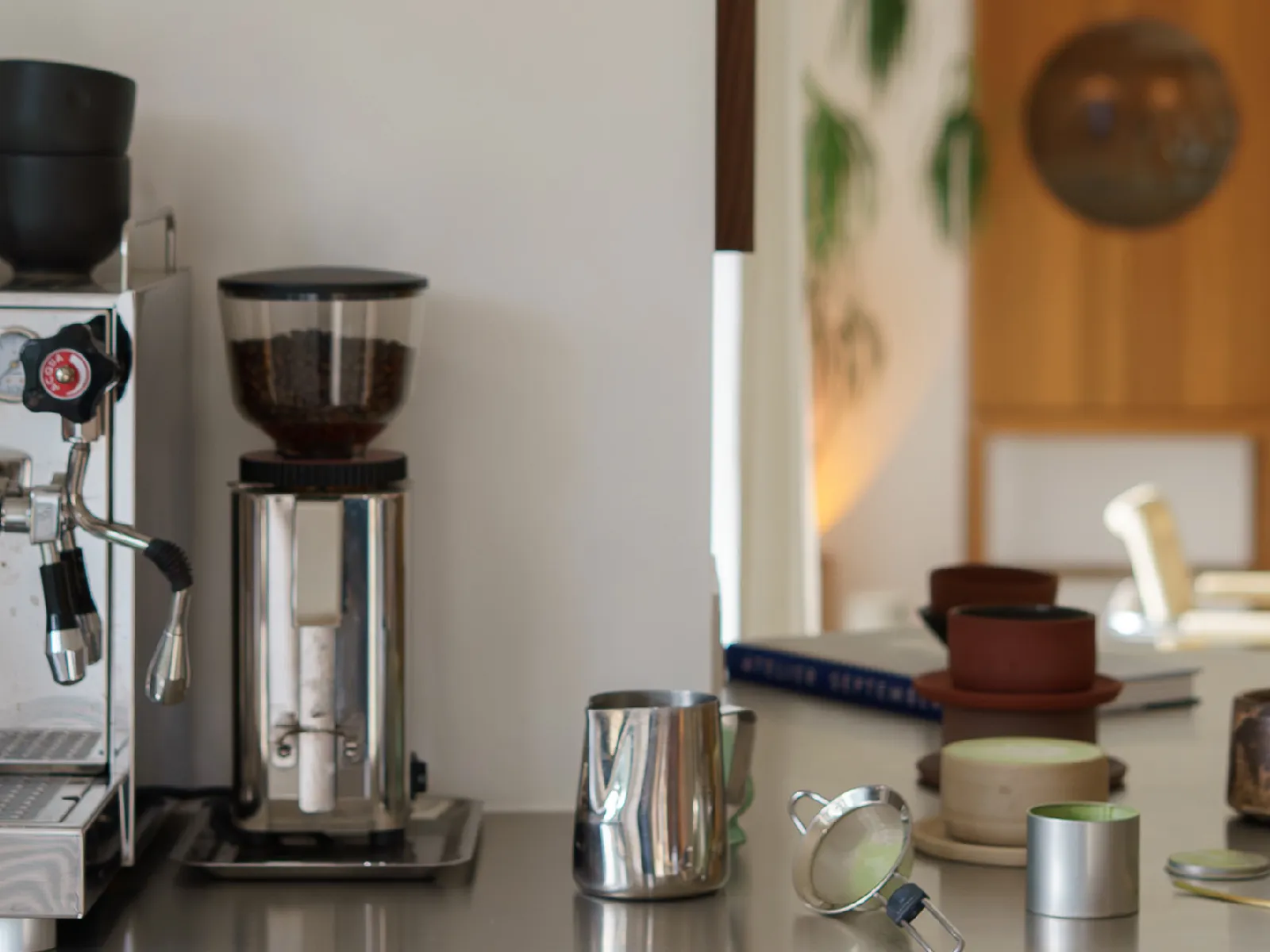A coffee station with an espresso machine, grinder, and various matcha tea preparation items on a light-colored counter.