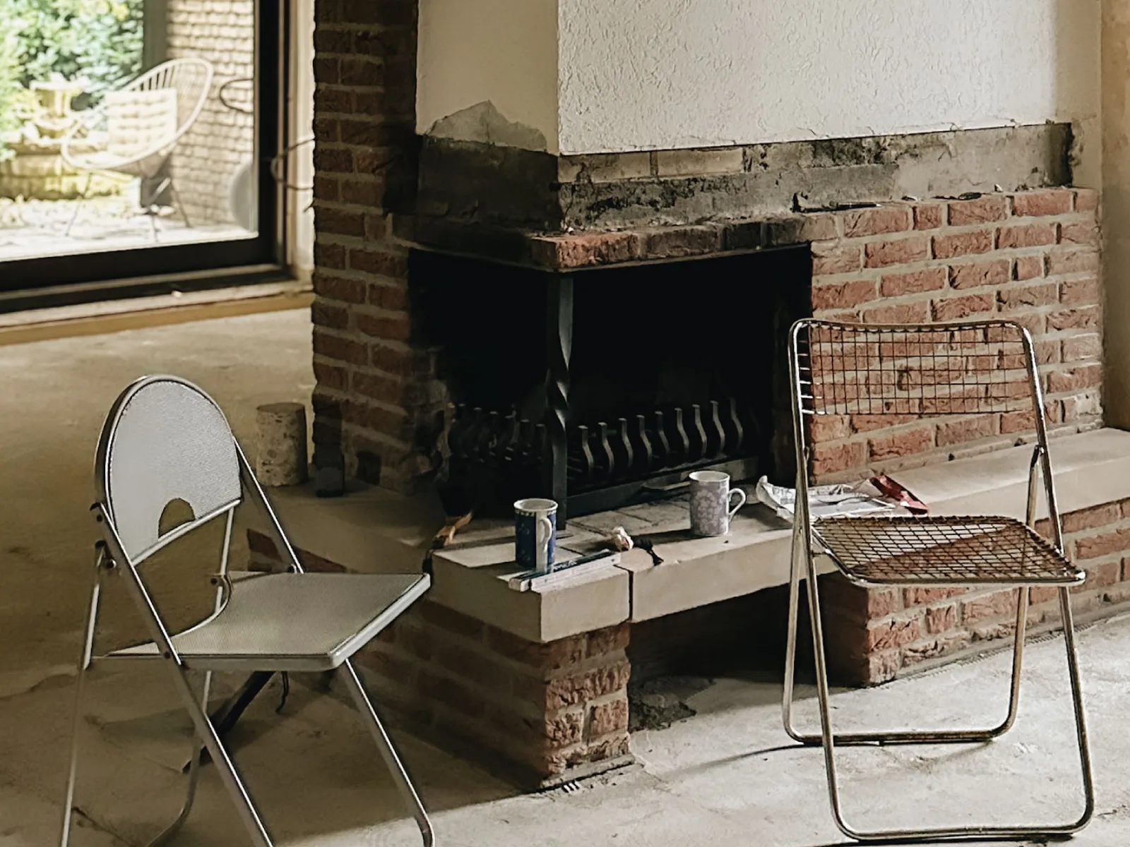 A room under renovation with a brick fireplace, two chairs, and a view of a garden through a glass door.