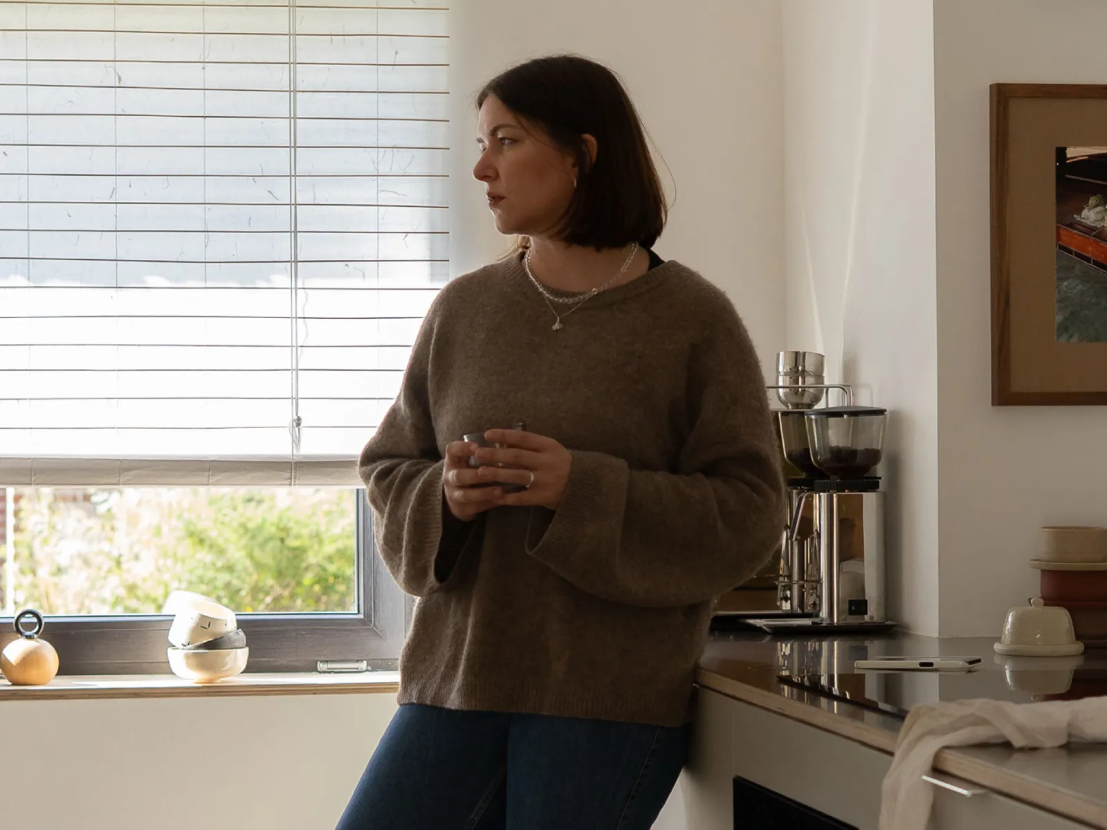 A woman in a brown sweater and jeans stands in a kitchen, holding a mug and looking out a window with blinds.
