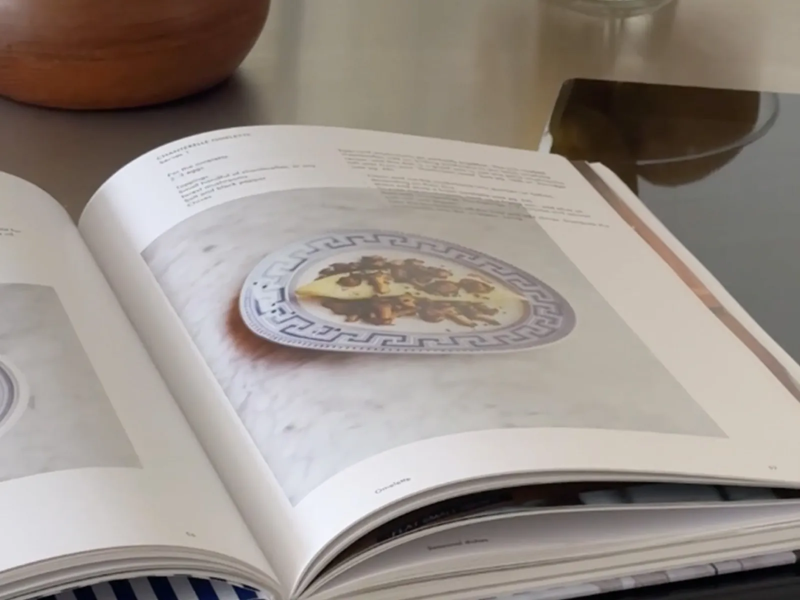 An open cookbook displays a recipe with an image of a dish on a decorative plate, resting on a reflective surface.