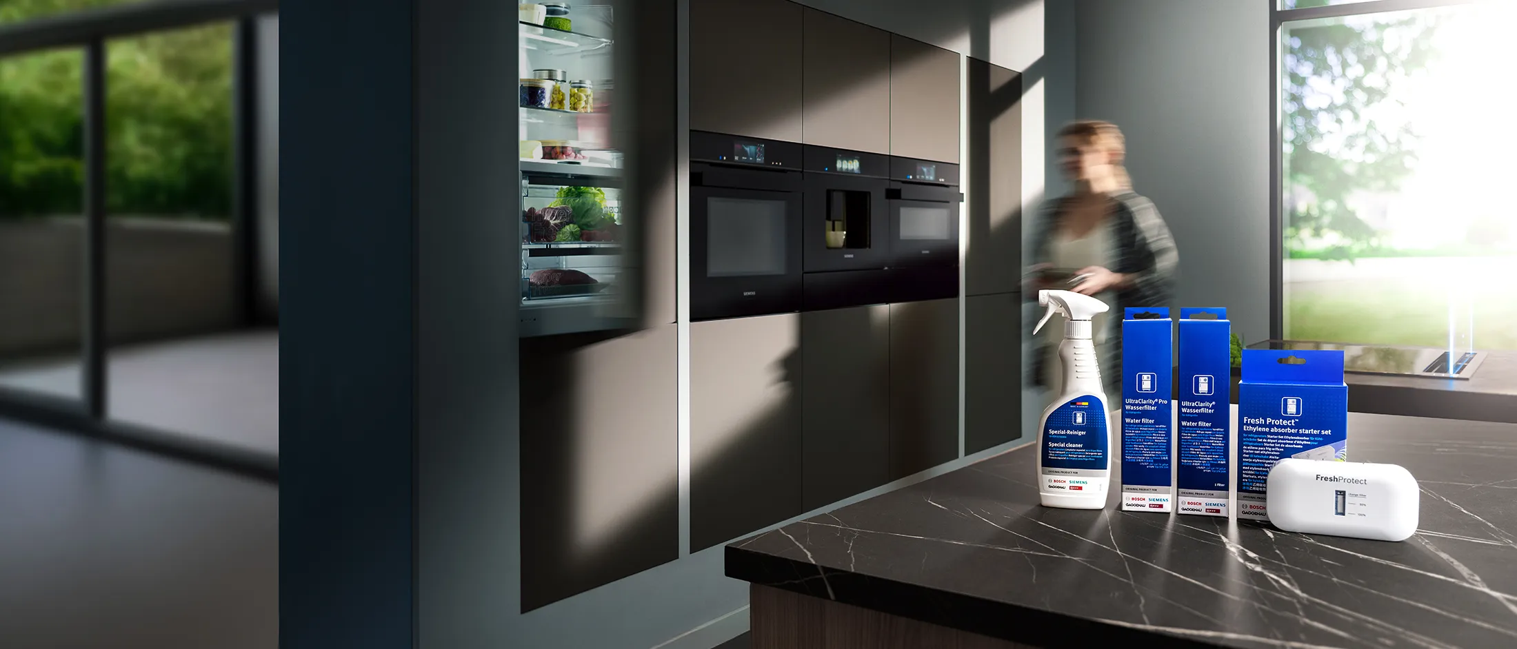 Modern kitchen with cleaning products on the counter and a blurred person moving near built-in ovens and an open refrigerator.