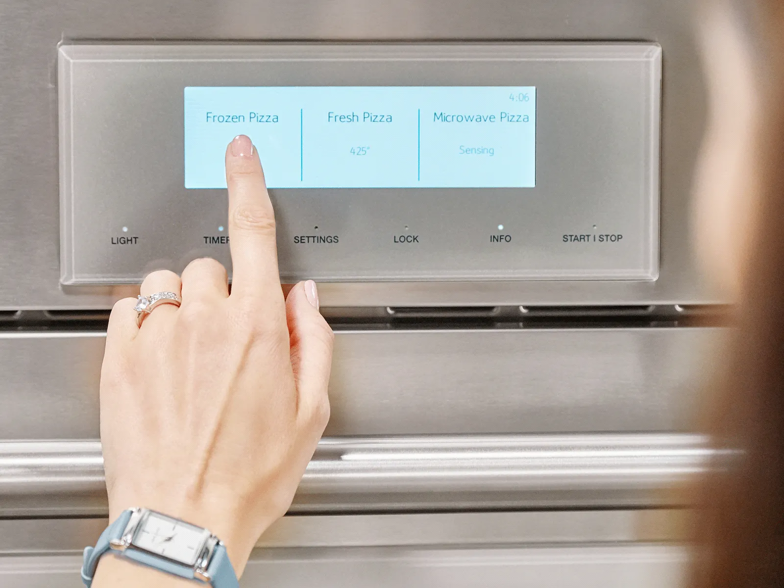 A hand selects 'Frozen Pizza' on a stainless steel oven touchscreen panel.