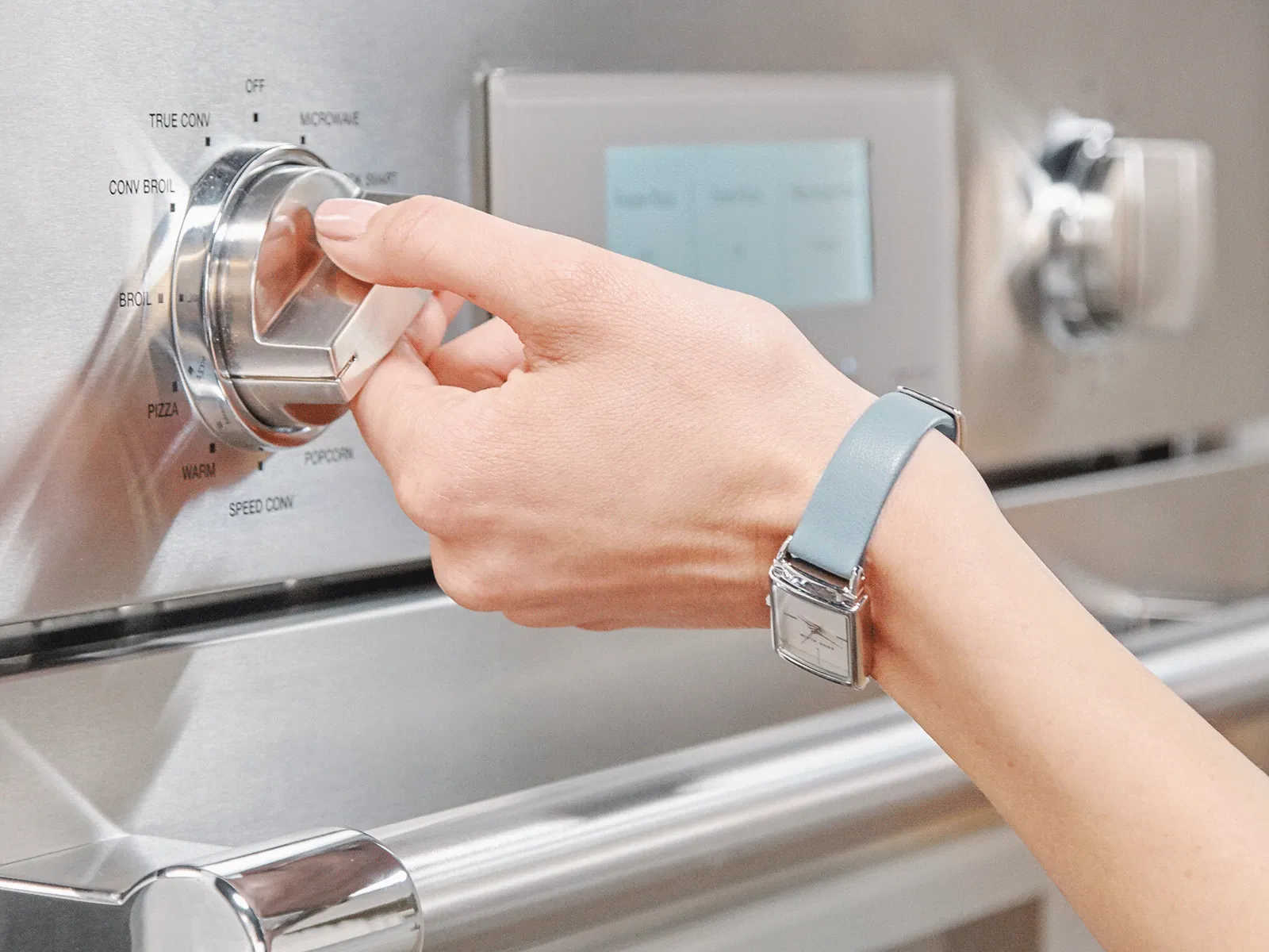 A hand adjusts the stainless steel oven dial to a cooking mode, with function options visible.