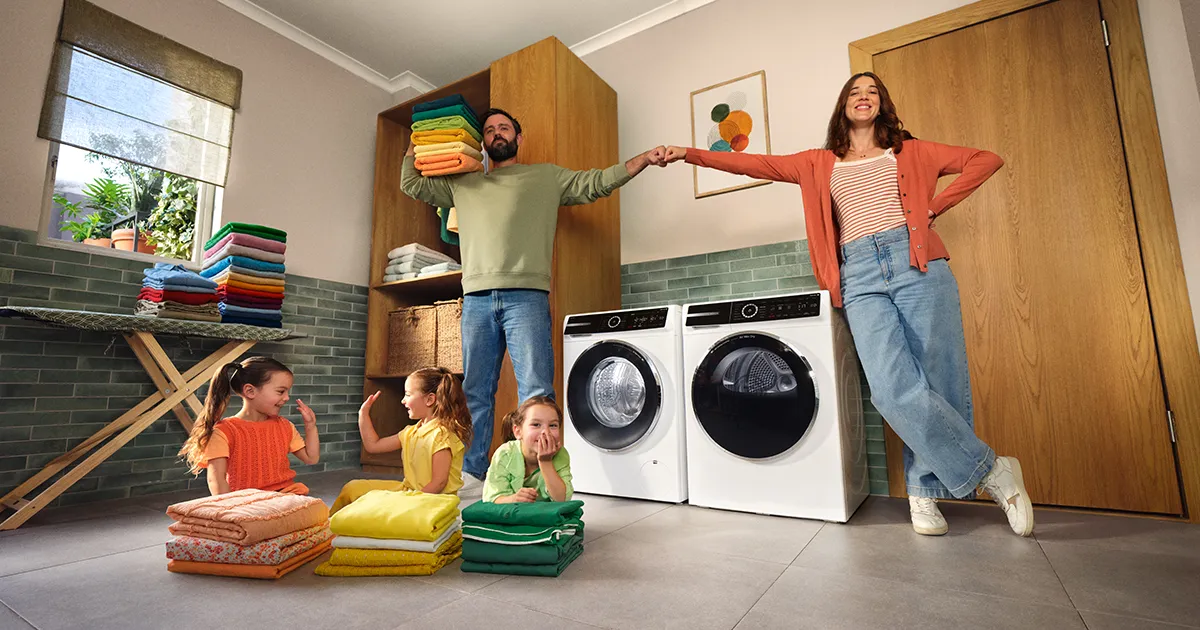Famille dans une buanderie avec deux enfants assis par terre pliant du linge coloré, un homme tenant des serviettes et une femme appuyée sur une machine à laver.
