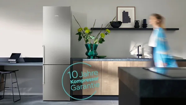 Moderne Küche mit silbernem Kühlschrank, Holzschränken, einer grünen Vase mit Blumen, einem schwarzen Regal mit Deko und einer vorbeigehenden Person, die unscharf abgebildet ist.