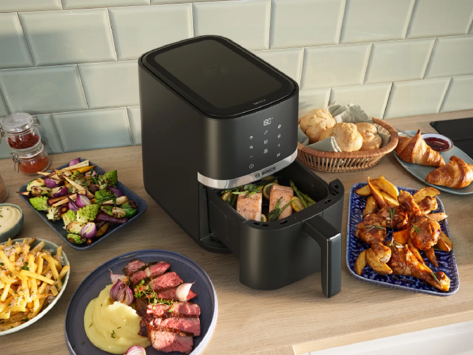 Bosch air fryer on a kitchen counter surrounded by various cooked dishes including steak, fries, salad, bread, and chicken wings.