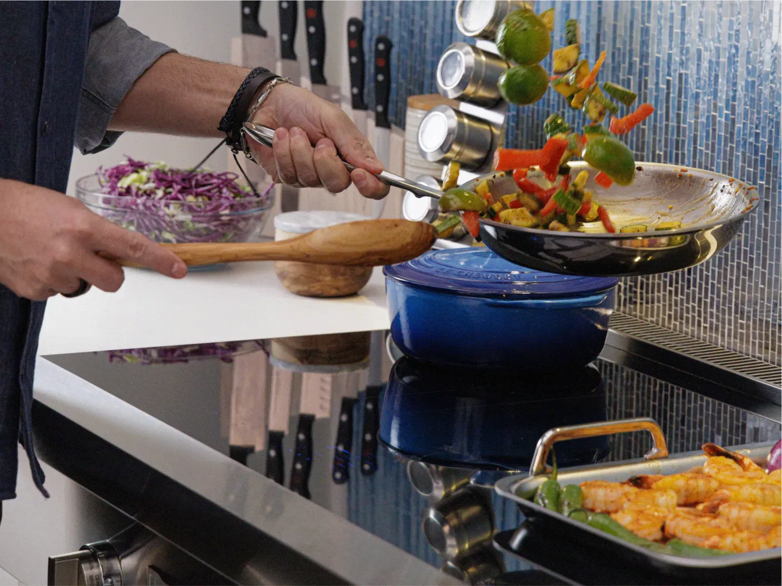 Person sautéing mixed vegetables in a pan over a stovetop with cooked shrimp in a tray nearby in a modern kitchen.