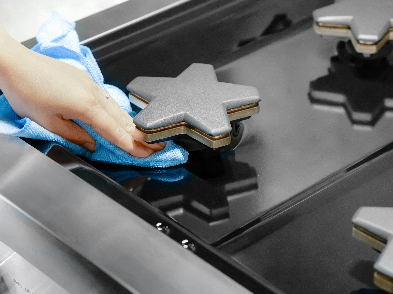 Hand cleaning a black gas stove with a blue cloth, focusing on a star-shaped burner cover.
