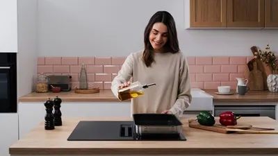 alt-Woman pouring cooking oil into a frying pan on a kitchen island with an induction cooktop.