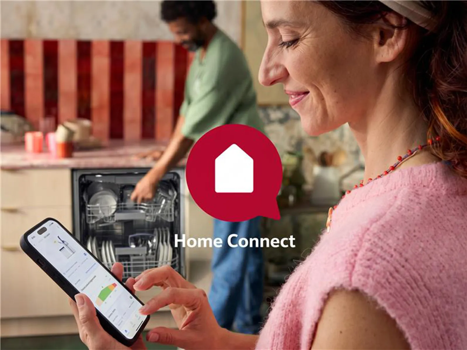 A nő a telefonján a Home Connect alkalmazást használja, miközben a férfi betölti a mosogatógépet.