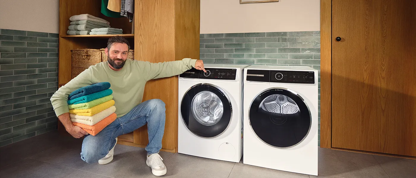 Man with folded colorful towels adjusting settings on a white washing machine next to a dryer in a modern laundry room.