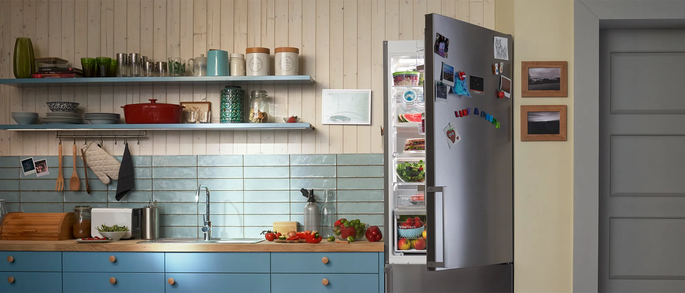 In a kitchen. Bosch refrigerator with an opendoor, filled with fresh food. Letter magnets on the door read "Like a Bosch.”