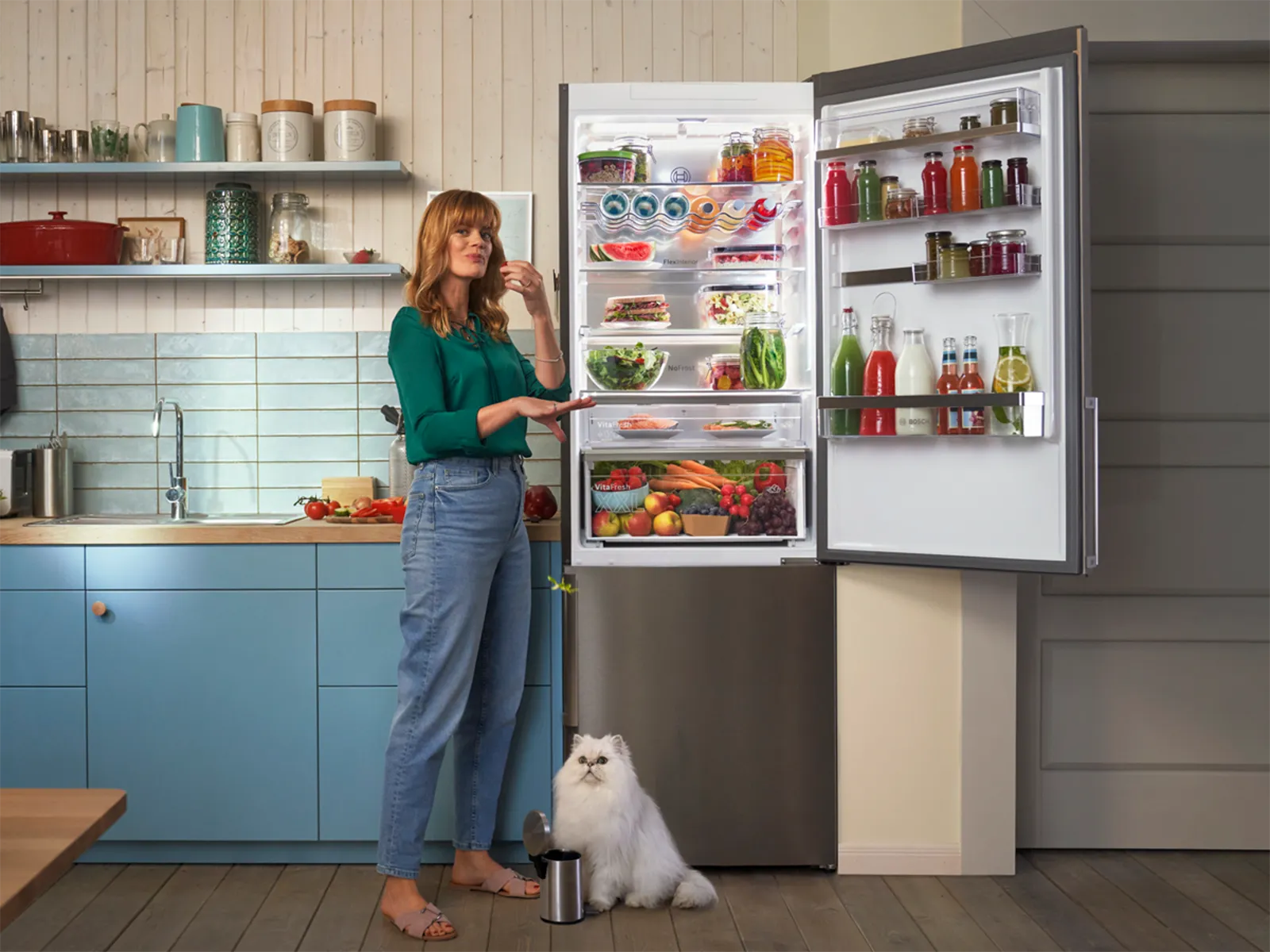 Woman standing in a kitchen, eating near an open fridge filled with food, while a white cat sits beside a small trash bin on the floor.