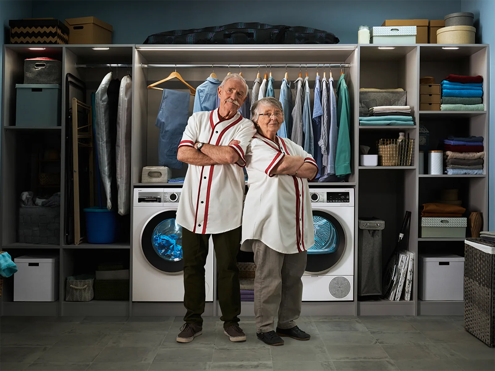 Two people in matching shirts stand confidently in a modern laundry room with washer, dryer, and organized shelves behind them.