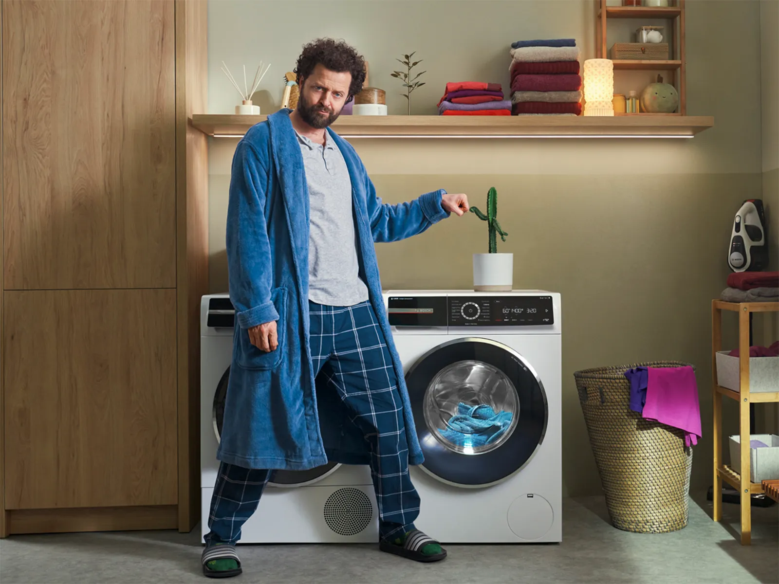 Person in blue robe and pajamas standing beside a washing machine in a laundry room with folded towels and a cactus plant on top.