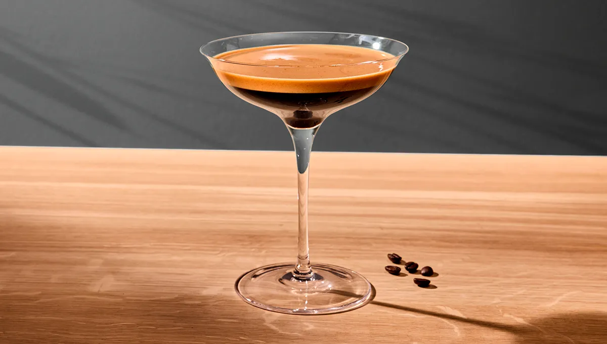 Espresso Martini na svetlej drevenej doske s niekoľkými kávovými zrnami vedľa neho.