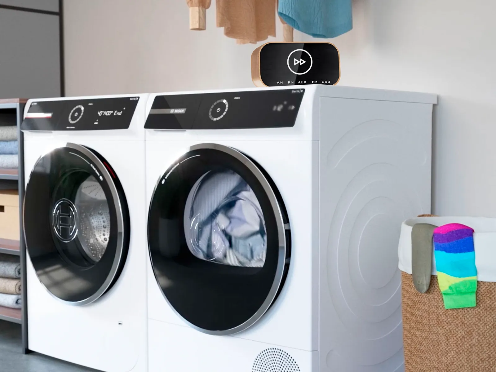 Lavatrice e asciugatrice Bosch bianche a carica frontale, con dei panni dentro l'asciugatrice, uno smart speaker appoggiato sopra e un cesto del bucato con roba colorata lì vicino.