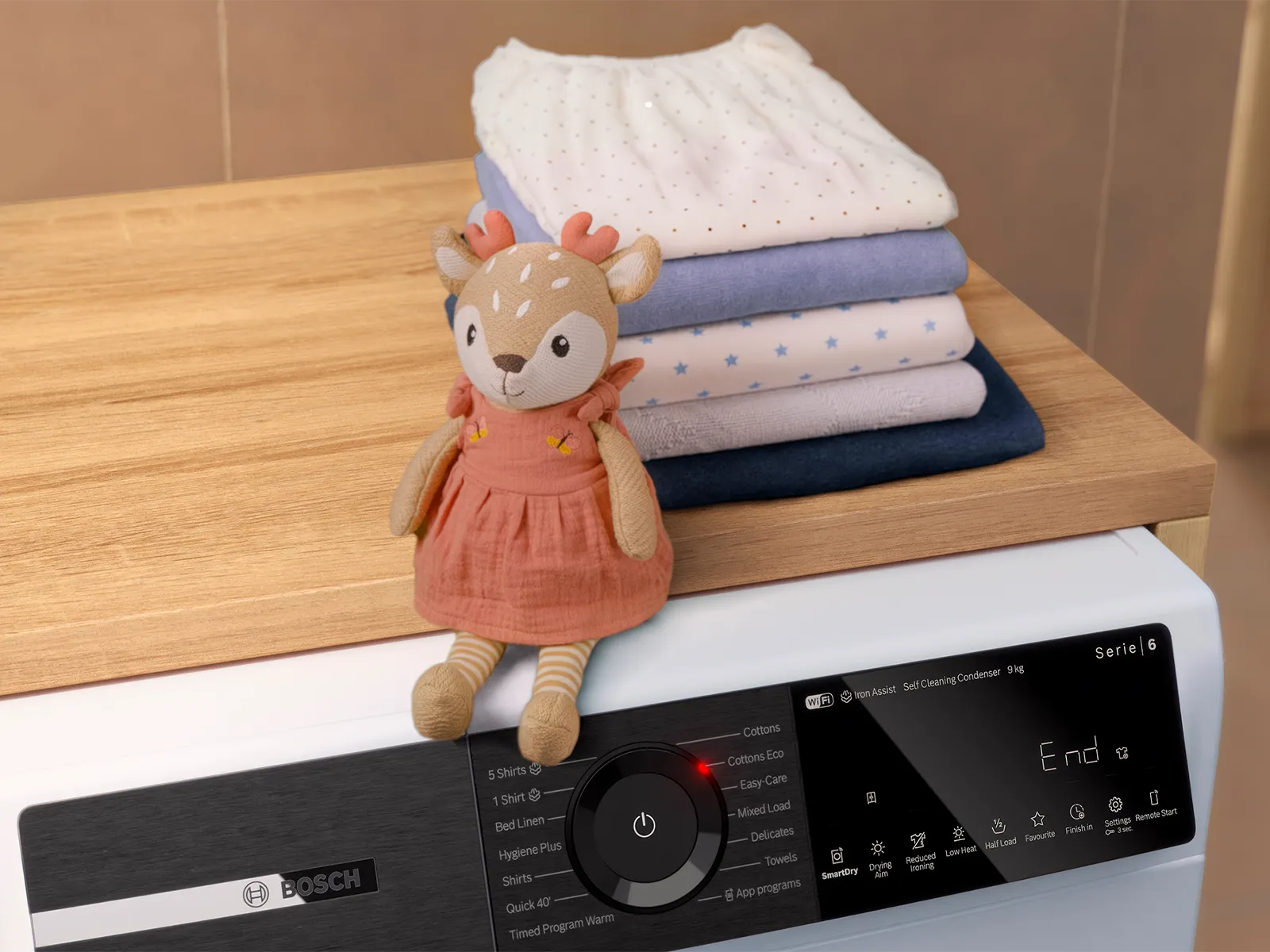 Un cerbiatto di peluche con un vestitino è seduto su una lavatrice. Dietro, su un piano di legno, c'è una pila di vestiti per neonati, tutti piegati