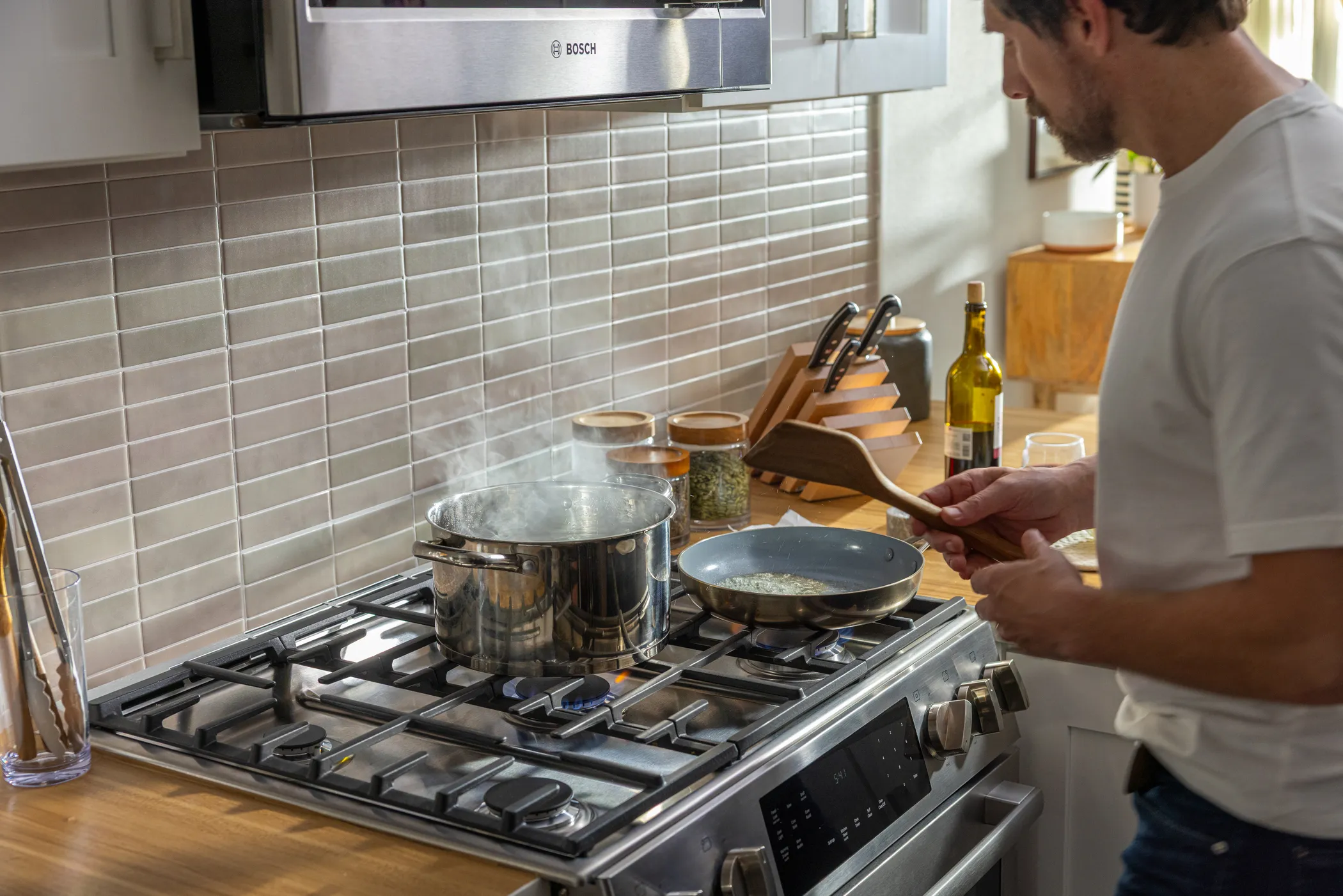 Woman smiling while pouring oil into a pan on an induction stove in a modern kitchen with wooden countertops and pink tile backsplash.