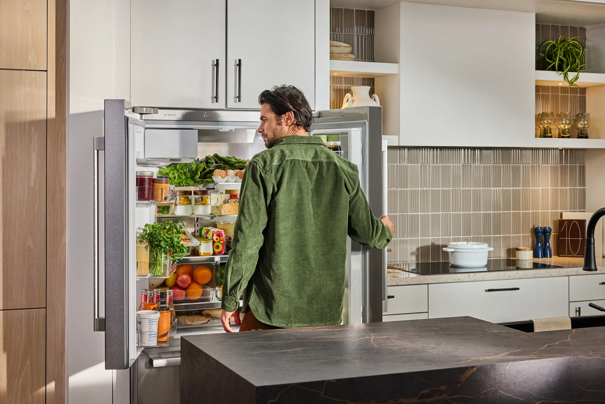 Man in green shirt opens a Bosch stainless steel fridge in a modern kitchen filled with fruits and vegetables.