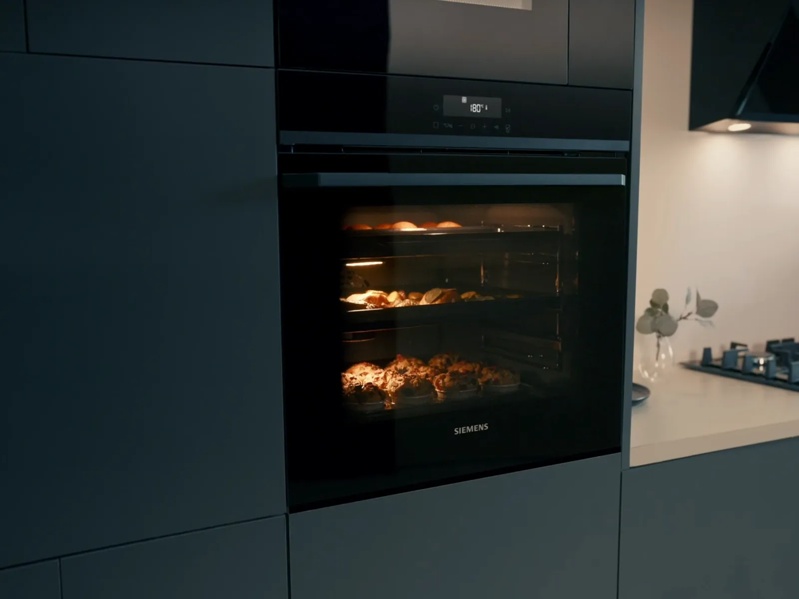 Black built-in oven with food cooking inside, integrated into dark gray kitchen cabinets. Gas hob visible.