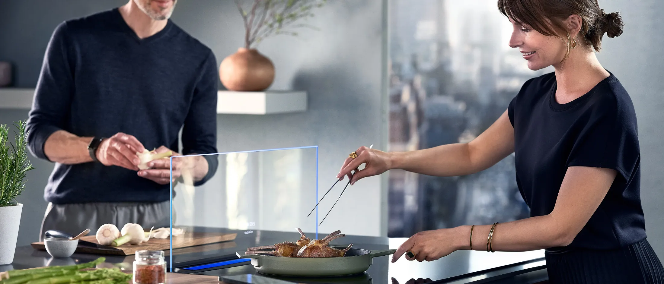 Två personer som lagar mat tillsammans i ett modernt kök, en vänder kött i en panna medan den andra förbereder svamp på en skärbräda.