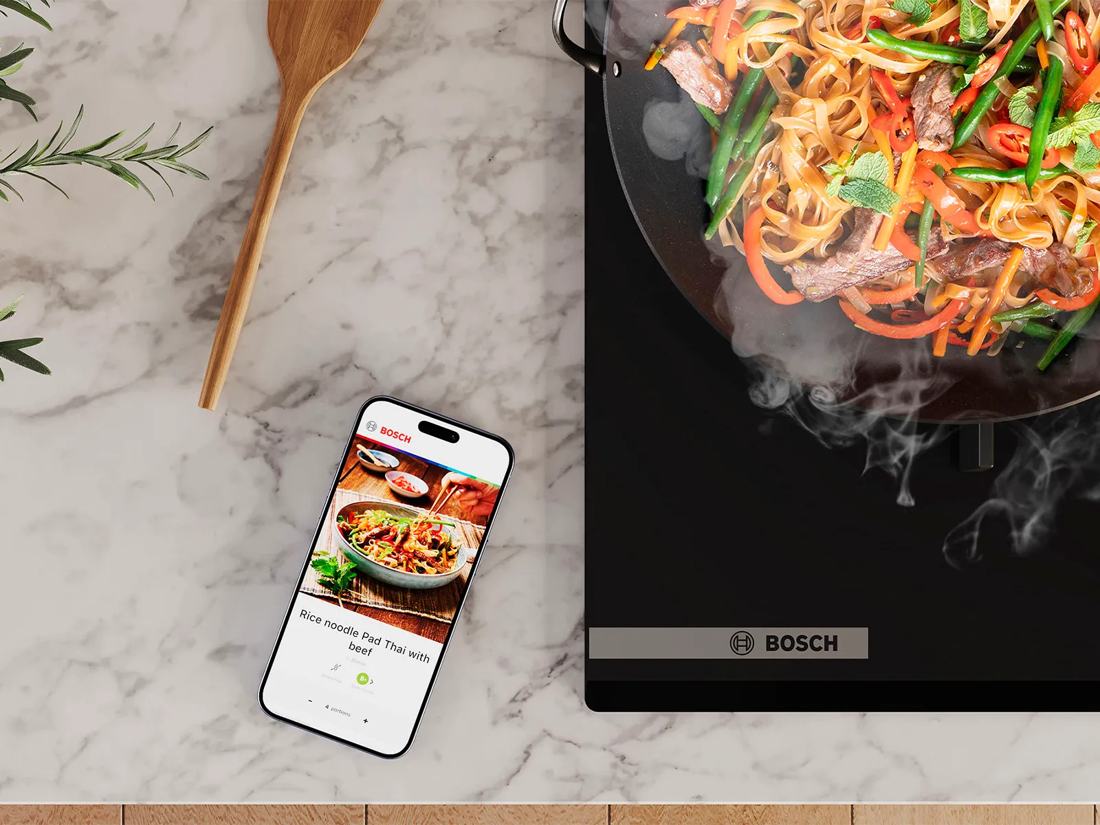 Smartphone che mostra una ricetta accanto a un wok fumante con noodle saltati, verdure e manzo su un piano di lavoro in marmo con una pianta e un cucchiaio di legno.