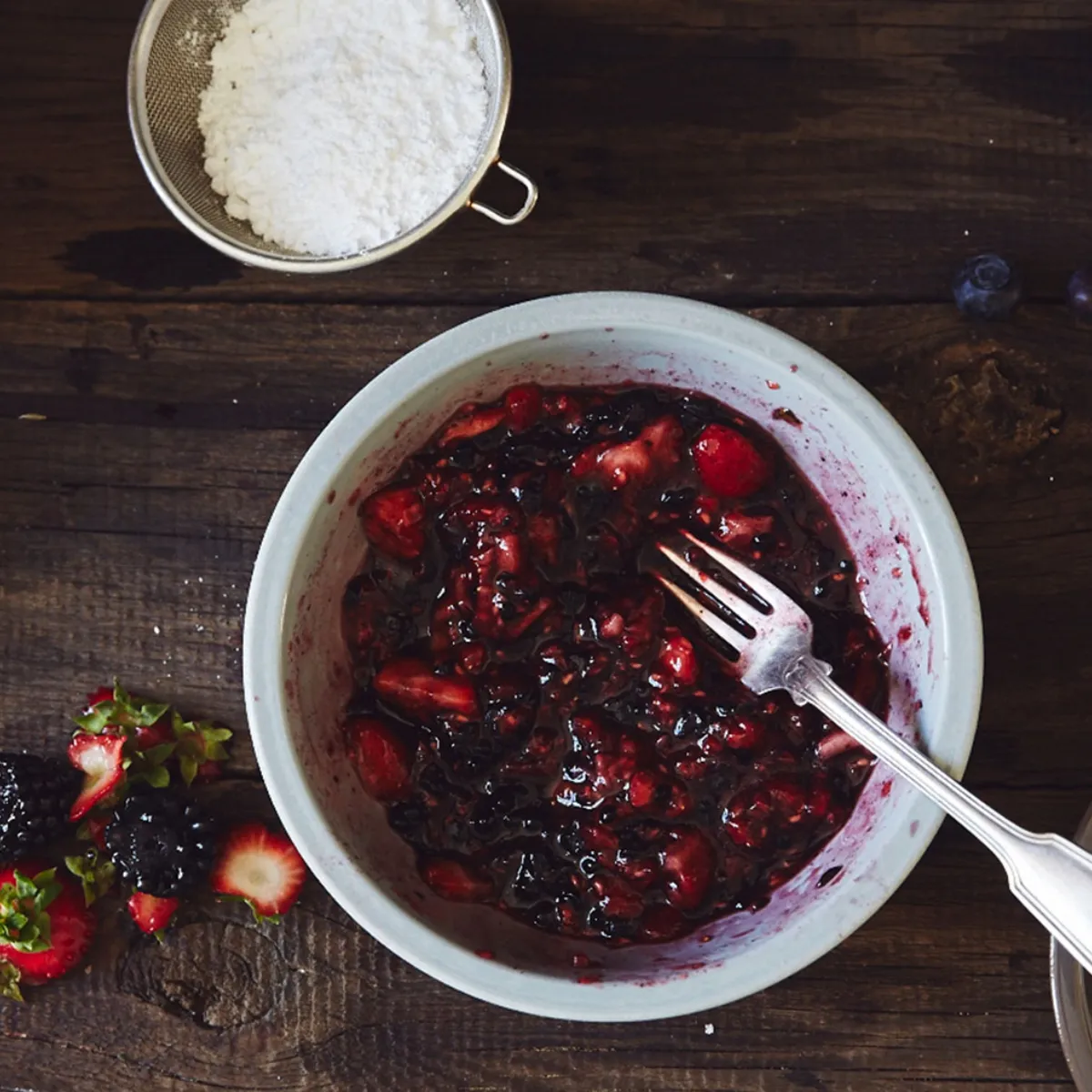 An overhead view of a bowl of mixed berries (strawberries, blackberries, raspberries) mashed with a fork, surrounded by fresh berries, powdered sugar, and wooden popsicle sticks on a rustic wooden table.