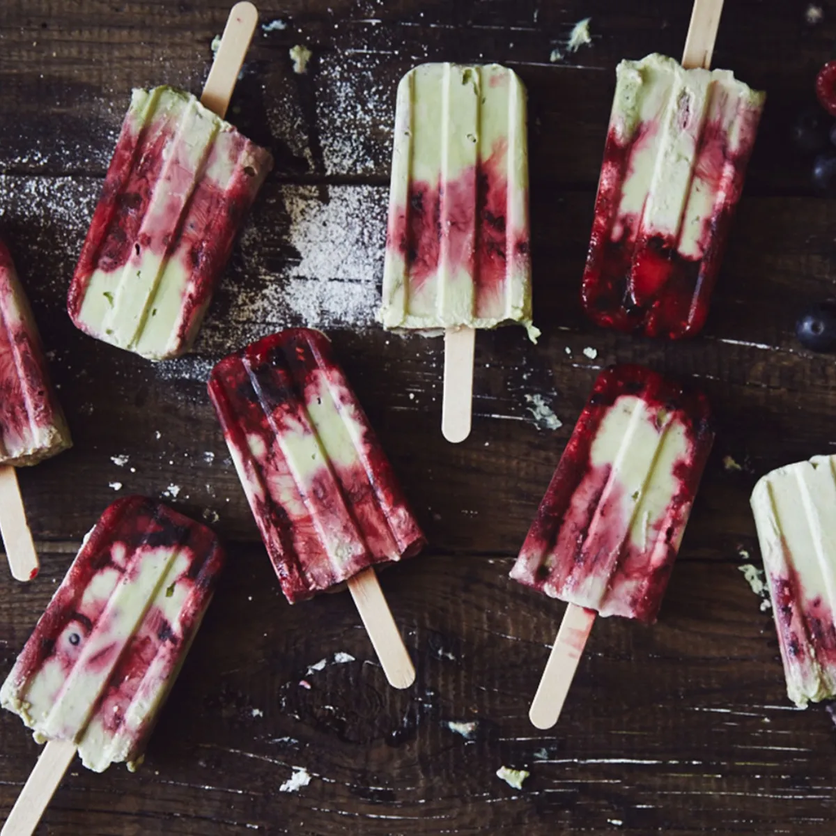 Several homemade green and red fruit popsicles with wooden sticks are arranged on a rustic wooden surface, surrounded by fresh blueberries and raspberries.