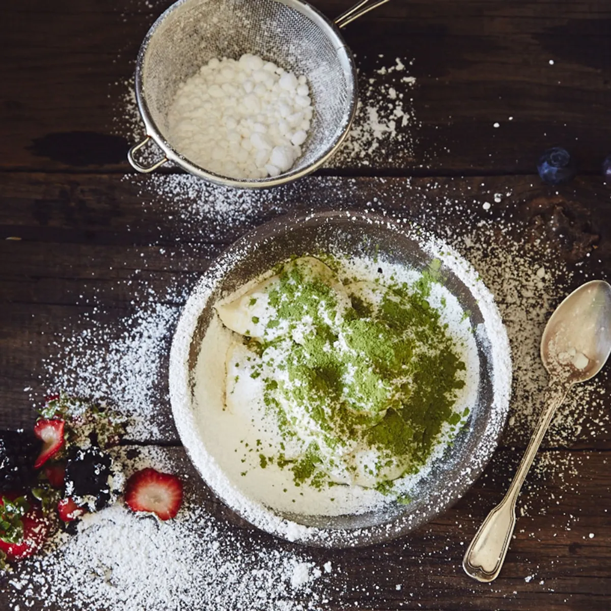 An overhead view of a bowl of whipped cream topped with green matcha powder, surrounded by powdered sugar, fresh berries, and a spoon on a rustic wooden table.