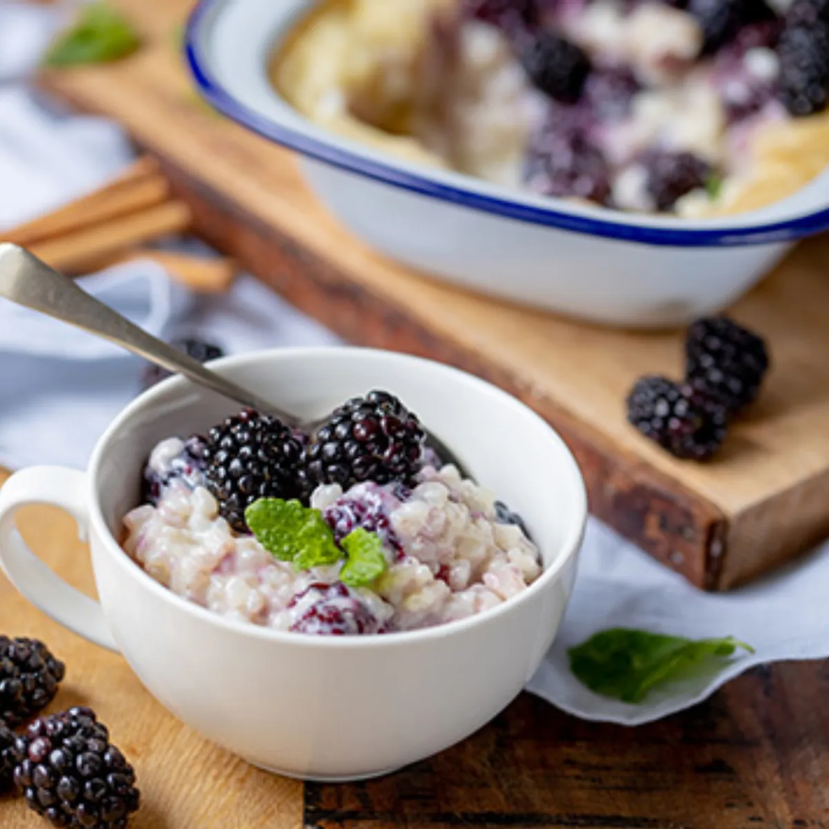 A white mug of creamy rice pudding with fresh blackberries and mint, next to a baking dish of blackberry cobbler and cinnamon sticks on a wooden board.