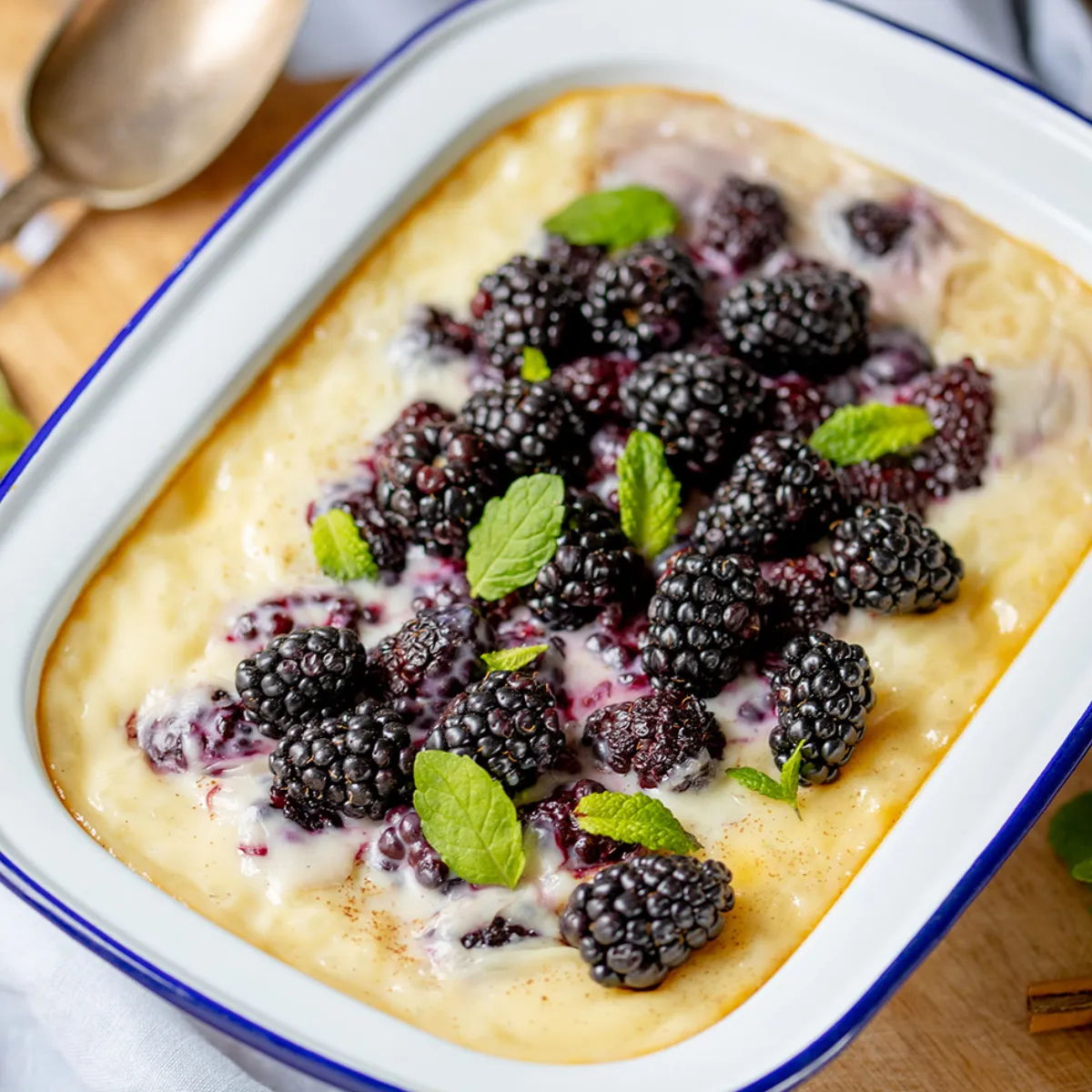 A white enamel baking dish filled with a creamy dessert topped with fresh blackberries, sitting on a wooden table.