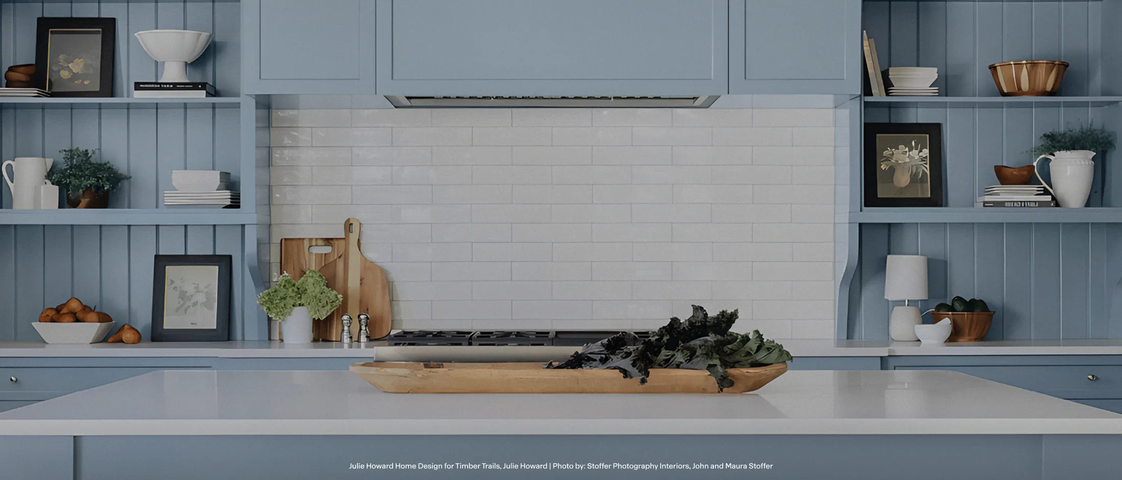 Modern blue kitchen with white countertop island, open shelves, wooden cutting boards, decor items, and a leafy centerpiece.