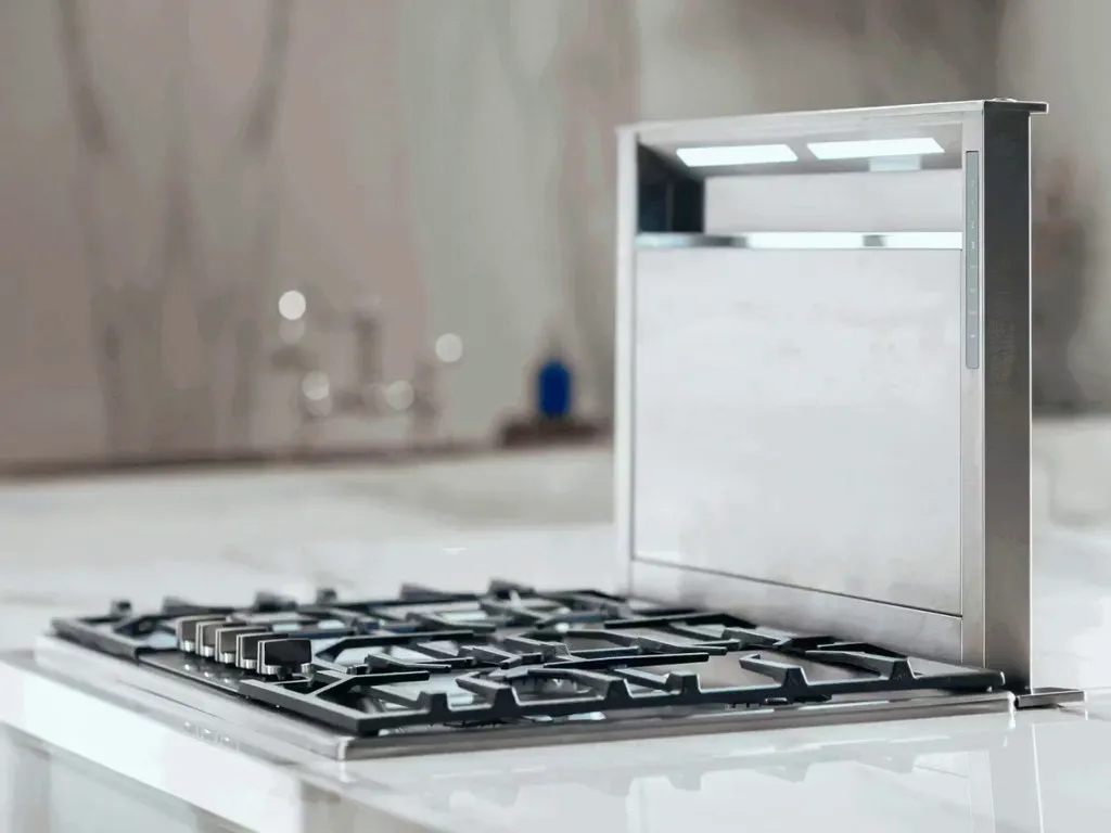 Stainless steel gas stovetop with multiple burners and integrated pop-up vent on a white marble countertop.