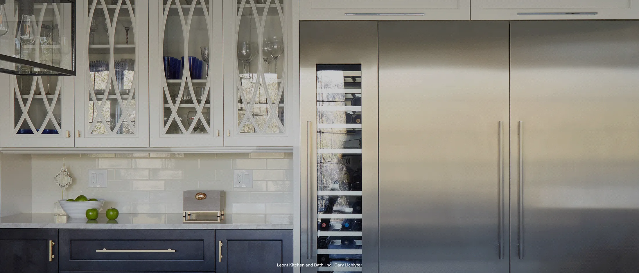 Built-in stainless steel refrigerator and wine column set in a modern kitchen, with glass-front cabinets, white tile backsplash, and countertop with fruit and kitchen accessories.