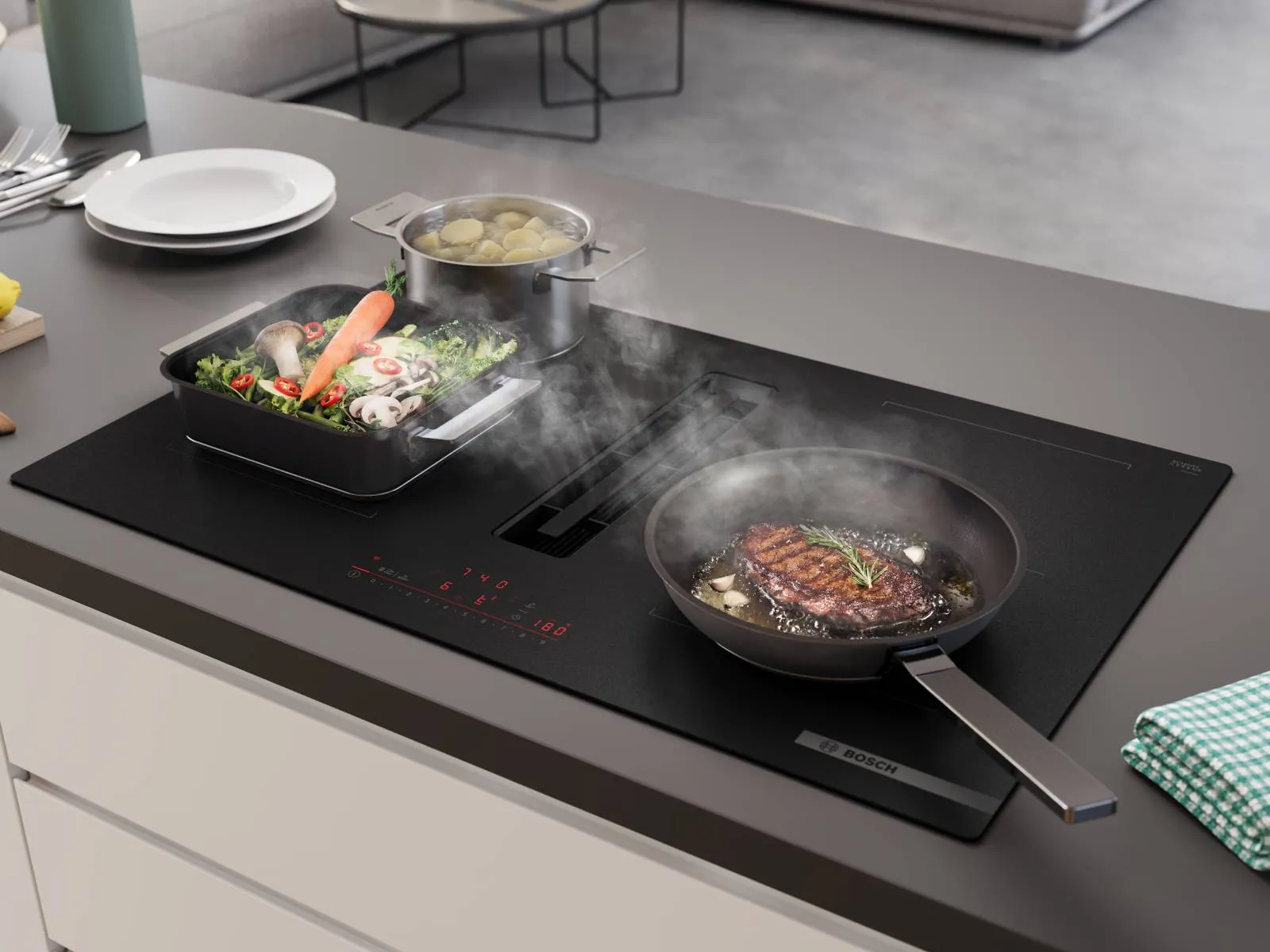 A black Bosch induction cooktop with an integrated downdraft extractor actively pulling steam from a pot of broccoli, set in a modern kitchen island.