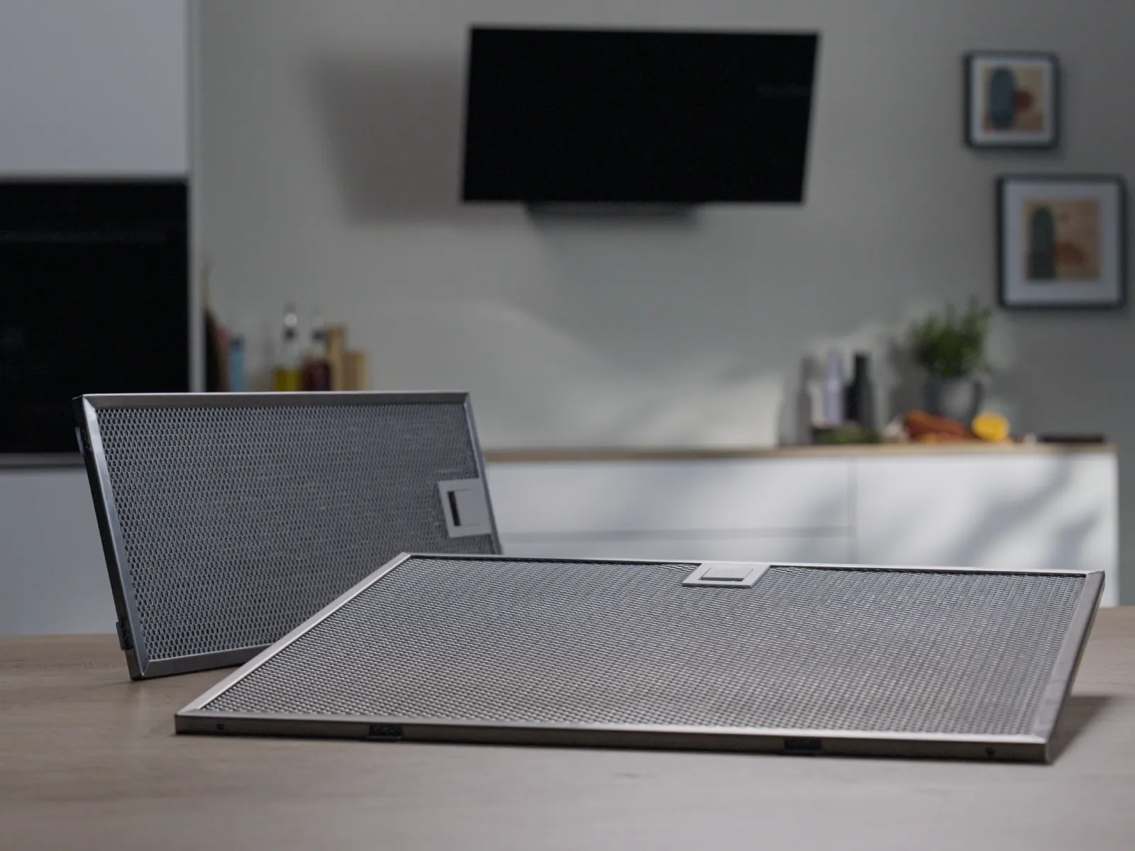 Två rektangulära metallnätfilter till köksfläkten placerade på ett träbord i en modern köksmiljö.