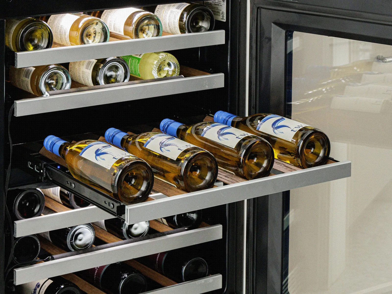 Open under-counter wine cooler with multiple wine bottles stored on sliding shelves in a modern kitchen setting.