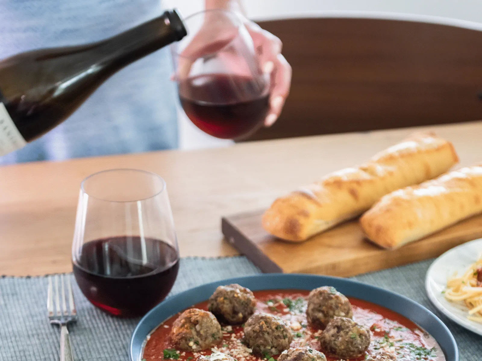 Person serving meatballs with spaghetti and bread on a wooden table, with glasses of red wine being poured.