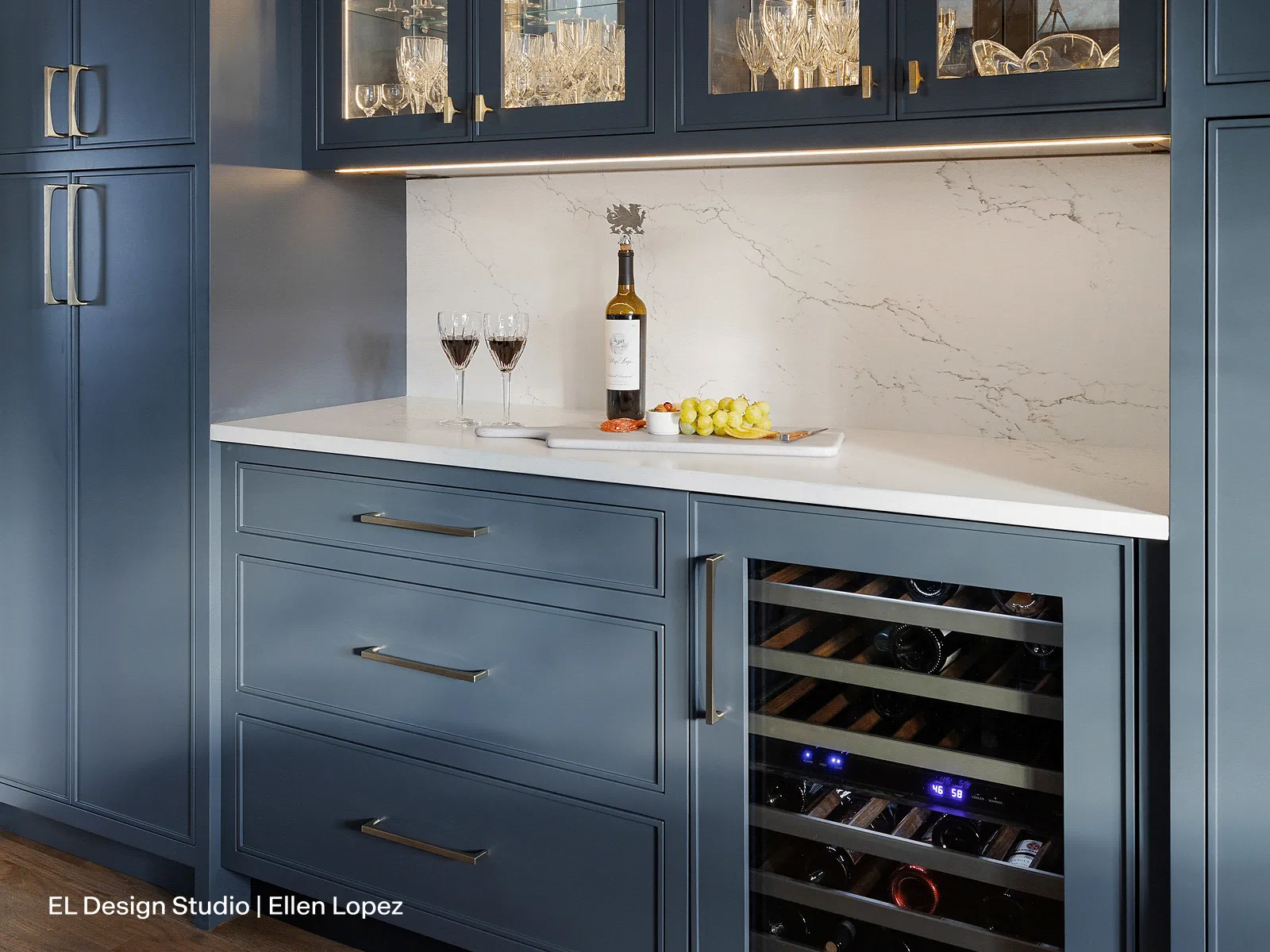 Modern blue kitchen cabinetry with glassware display, wine cooler, marble backsplash, and countertop holding wine glasses, bottle, and grapes.
