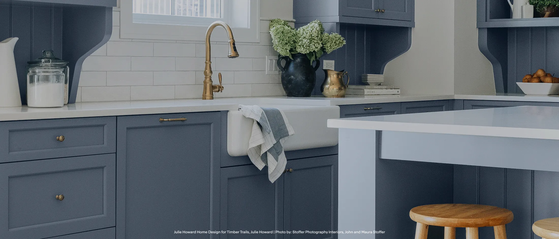 Modern kitchen with blue cabinets, white countertops, a brass faucet, a white dish towel, and a wooden stool by the island.