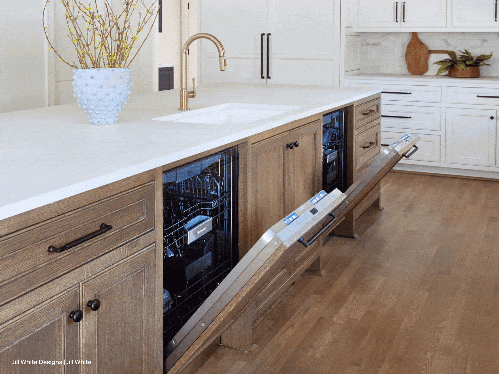 Modern kitchen featuring wooden cabinetry, open dishwasher, white countertop, gold faucet, and hardwood flooring with a decorative vase.